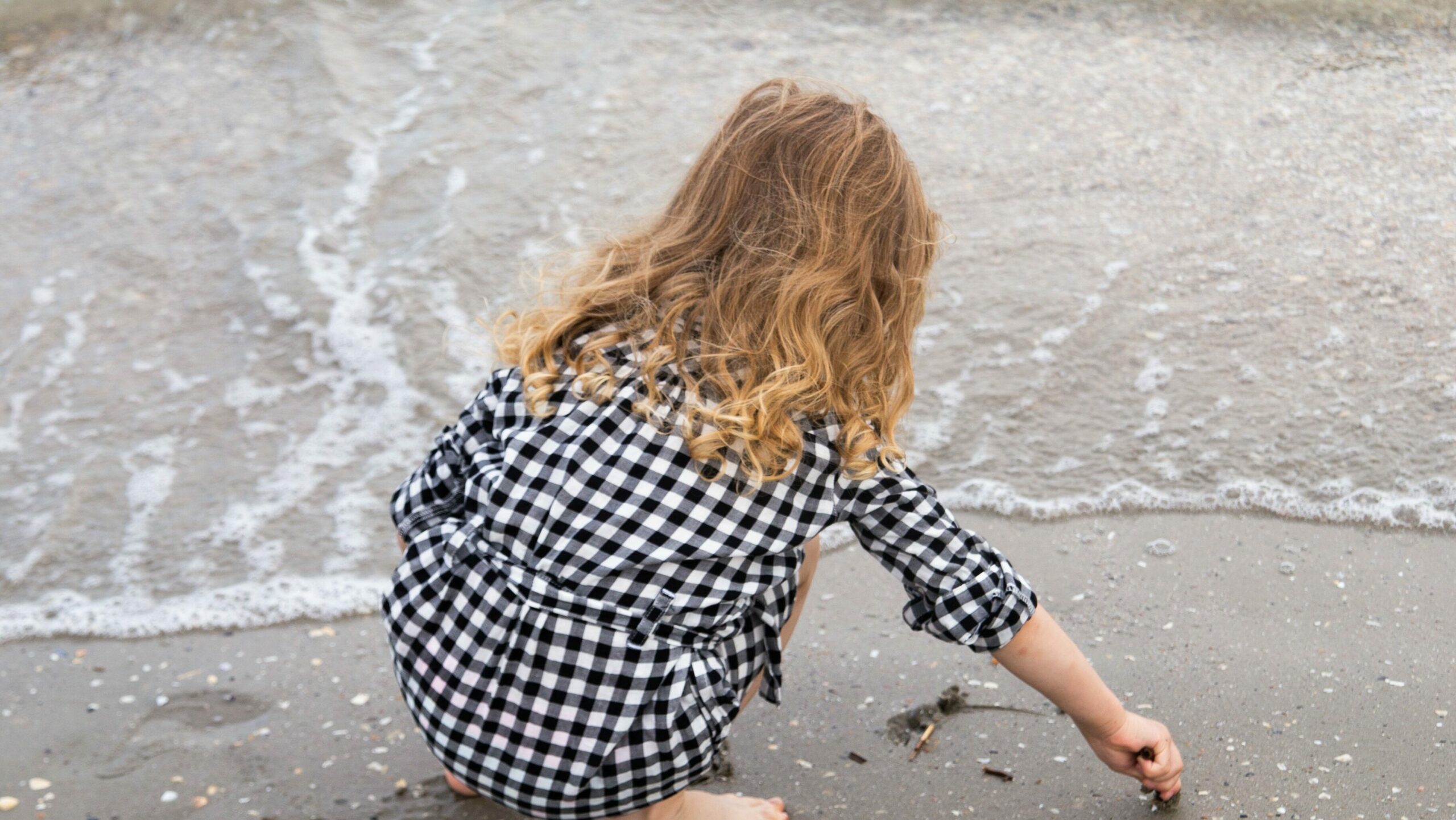 Une jeune fille de 5 ans accroupie au bord de l'eau à la plage, jouant avec le sable. nos prestations de services proposent également la garde d'enfant de plus de 3 ans.