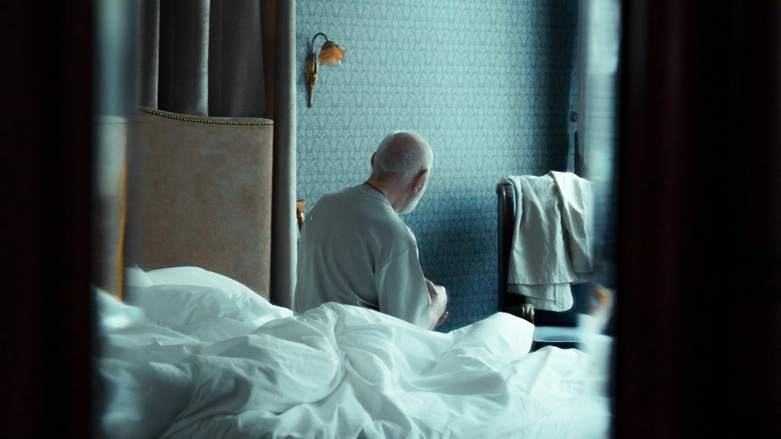 les premiers signes d'isolement chez une personne âgée ne se voient pas toujours. Ici il y a une image d'un homme âgé assis sur un lit défait, dos à la caméra, qui contemple le vide.