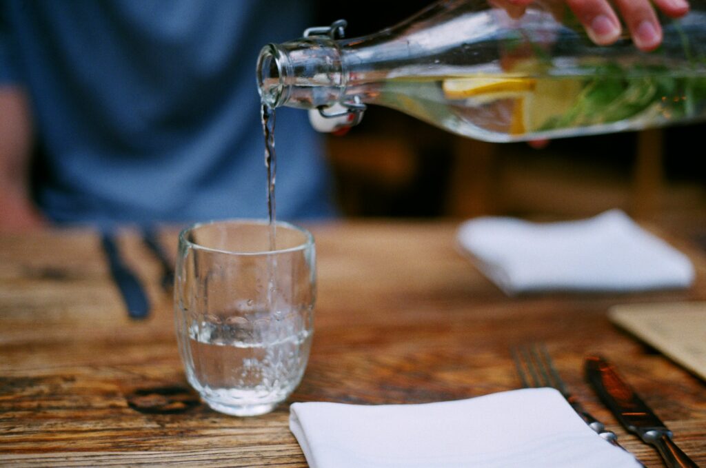 cette image représente un verre d'eau en train d'être rempli sur une table en bois. C'est comme cela que l'article dit qu'il faut prendre soin des personnes âgées pendant la canicule