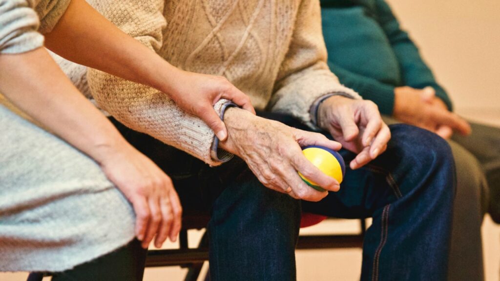 une femme tient le bras d'une femme plus âgées en signe de soutient. la personne agée tient une balle jaune et bleue dans la main. nos prestations de services proposent des accompagnements et sorties des personnes âgées et en besoin.