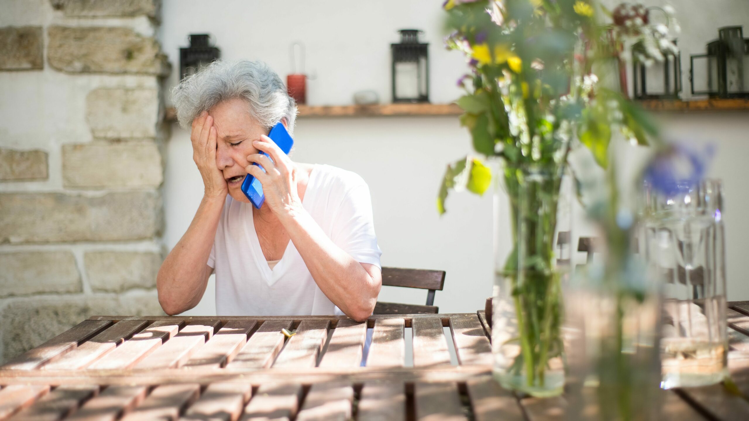 sur cette image nous voyons une dame âgée au téléphone assise à sa terasse sur une table en bois, sûrement dans son jardin. Elle a l'air en détresse.
Cette personne ne montre pas les premiers signes d'isolement chez une personne âgée mais semble tout de même en grande détresse